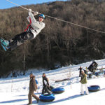 Snow tubers at Hawksnest in North Carolina watch as someone ziplines above the snow tubing lanes.